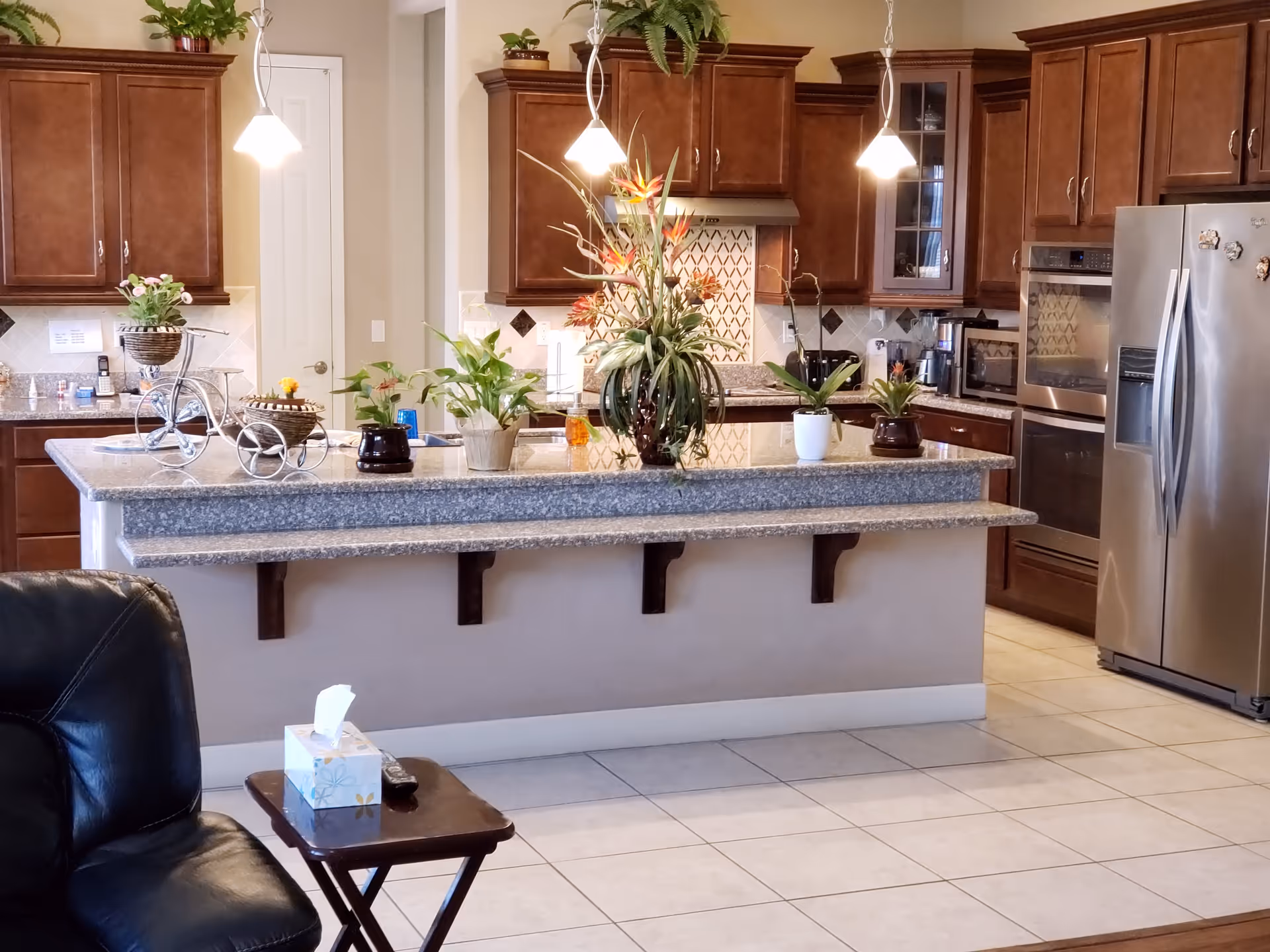 A modern kitchen with brown wooden cabinets, stainless steel refrigerator, oven, and microwave. A large granite countertop island is decorated with several potted plants and flowers. Three pendant lights hang above the island. In the foreground, there is a black leather chair and a small wooden side table with a tissue box and a remote control on it.