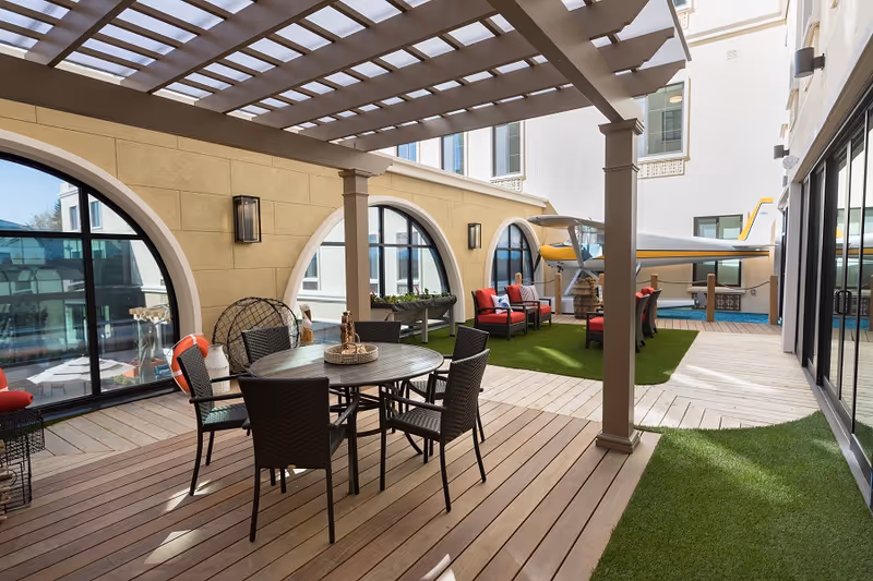 Outdoor patio area with a wooden deck and artificial grass, featuring a round table with six chairs under a pergola. There are red cushioned chairs and a small airplane displayed in the background near arched windows of a building.