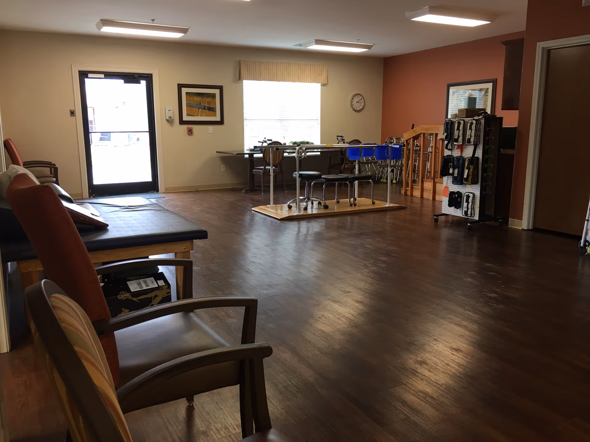 A spacious room with wooden flooring featuring several chairs along the left side, a padded bench, and a table with stools in the center. There is a window with blinds and a clock on the wall, a door with a window on the left, and a rack holding various medical or therapy equipment on the right side. The walls are painted beige and one wall is a reddish color.