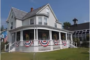 A large two-story white house with a wraparound porch decorated with red, white, and blue bunting. The house has multiple windows and a steep roof with a chimney. To the right, there is a smaller building with a cupola and a glass-covered walkway. The sky is clear and blue.