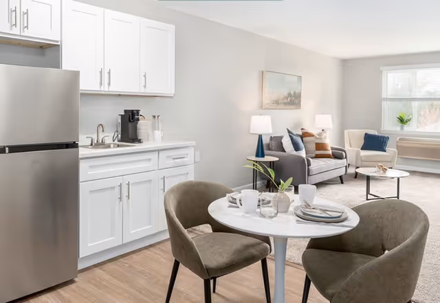 A bright and modern open-concept living space featuring a small kitchen area with white cabinets, a stainless steel refrigerator, and a coffee maker on the counter. In the foreground, there is a round white dining table set with plates, cups, and a small plant, surrounded by two upholstered chairs. In the background, a cozy living room area includes a gray sofa with decorative pillows, a white armchair, two table lamps, a round coffee table, and a large window letting in natural light.