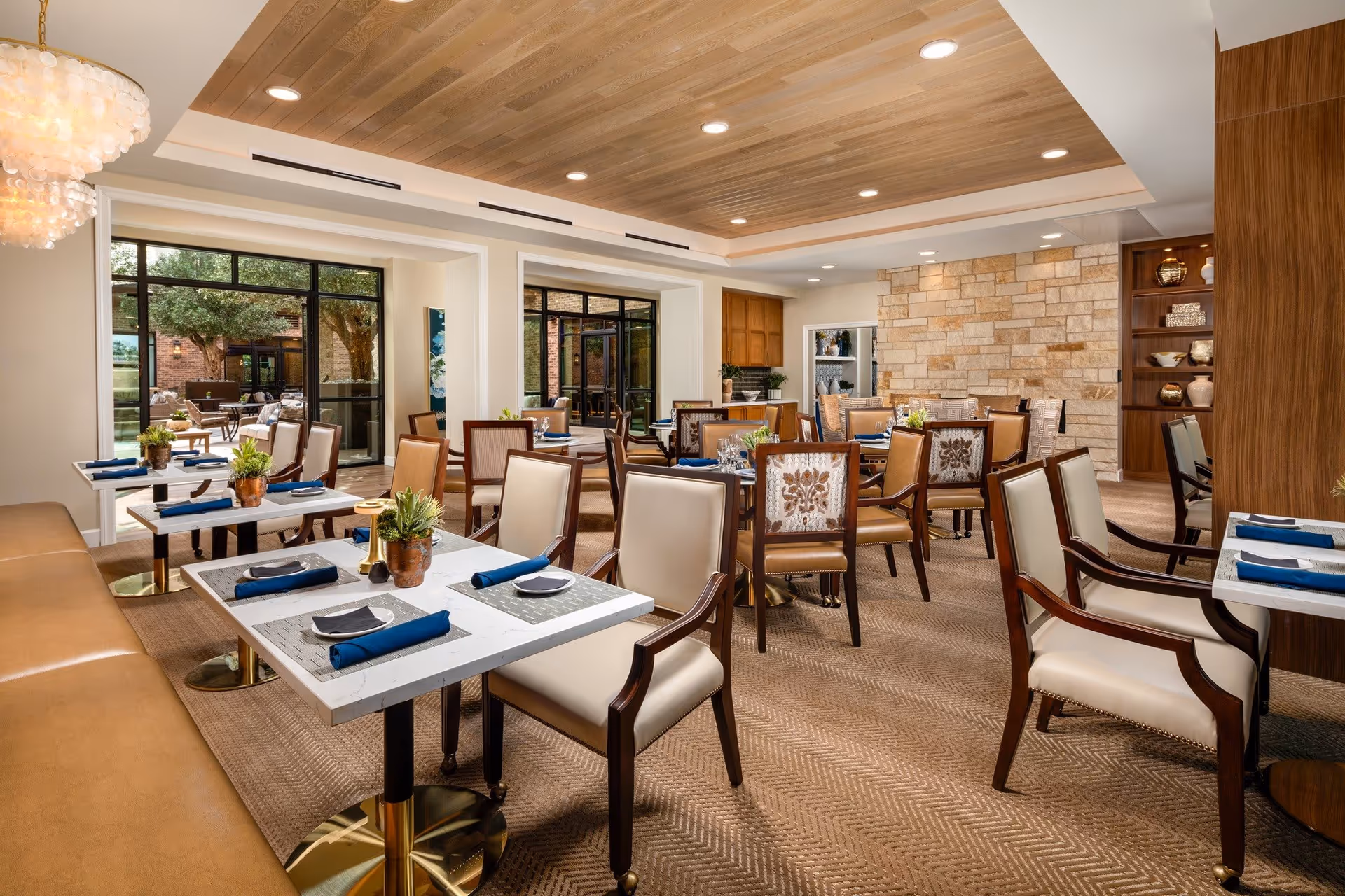 A well-lit dining room with multiple tables set with plates, napkins, and small potted plants. The room features beige upholstered chairs with wooden frames, a wooden ceiling with recessed lighting, a stone accent wall, and large windows looking out to an outdoor patio area with trees and additional seating.