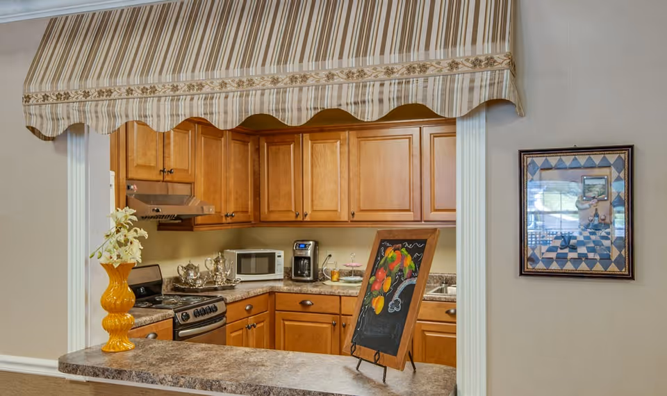 A cozy kitchen area with wooden cabinets, a stove, microwave, coffee maker, and a countertop with a yellow vase holding white flowers. A small chalkboard with colorful fruit drawings is displayed on the counter. There is a striped fabric awning above the kitchen pass-through window and a framed picture on the adjacent wall.