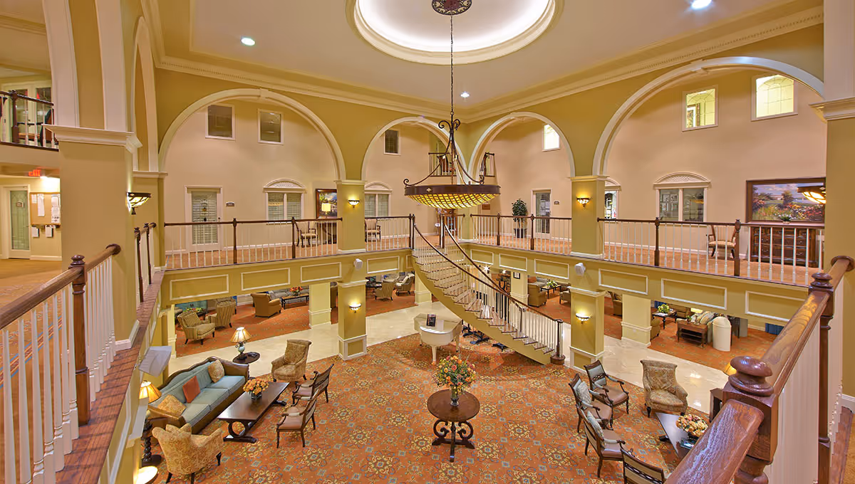 Spacious and elegant two-story common area in a senior living facility with a large central chandelier, a grand staircase, multiple seating areas with sofas and armchairs, and decorative arches along the upper level.