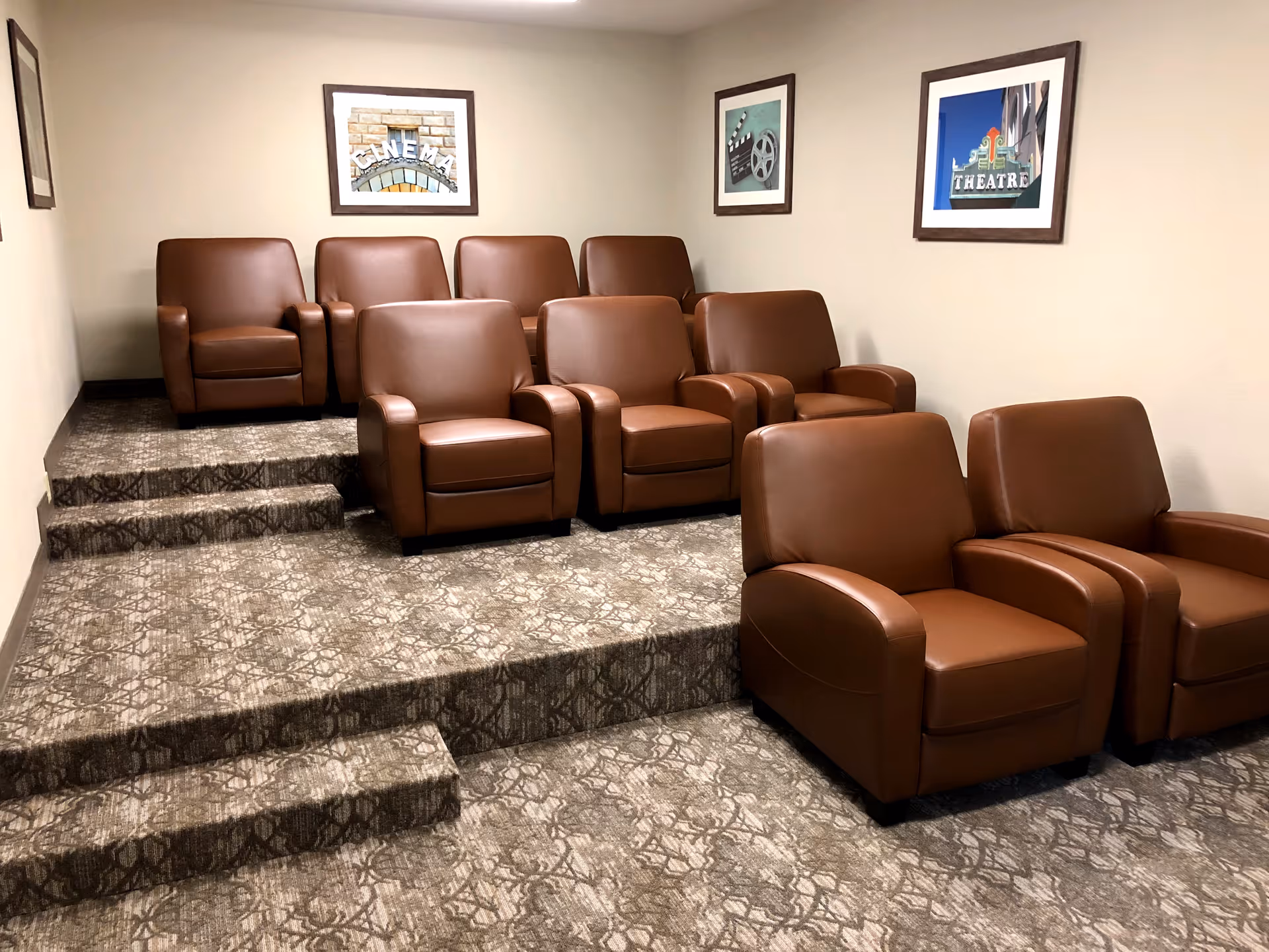 Small theater room with three rows of brown leather armchairs arranged on carpeted tiered platforms. The walls are beige and decorated with framed pictures related to cinema and theater.