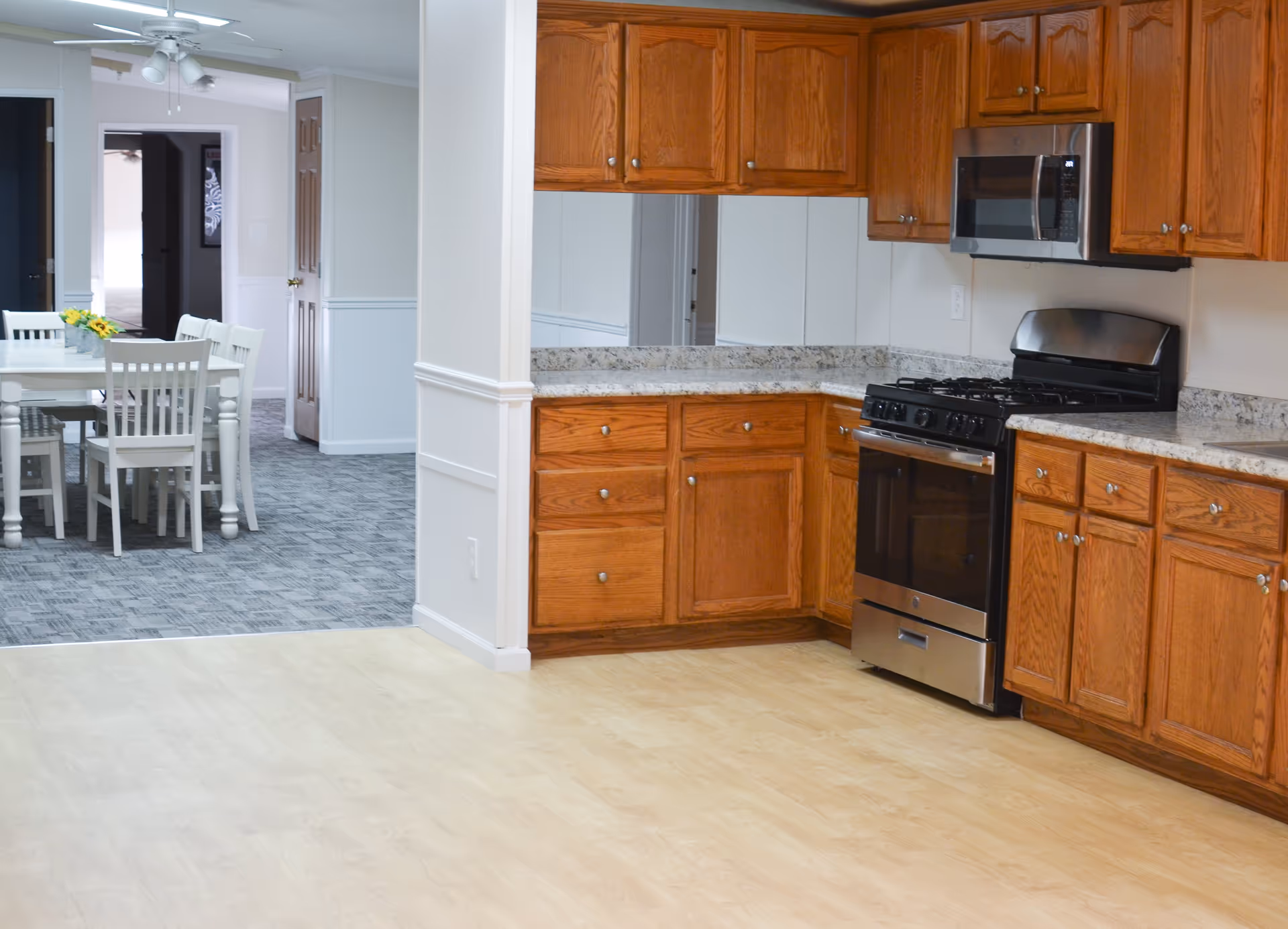 Interior view of a senior living facility kitchen with wooden cabinets, granite countertops, a stainless steel gas stove, and a microwave. Adjacent to the kitchen is a dining area with a white table and chairs, a ceiling fan, and carpeted flooring.