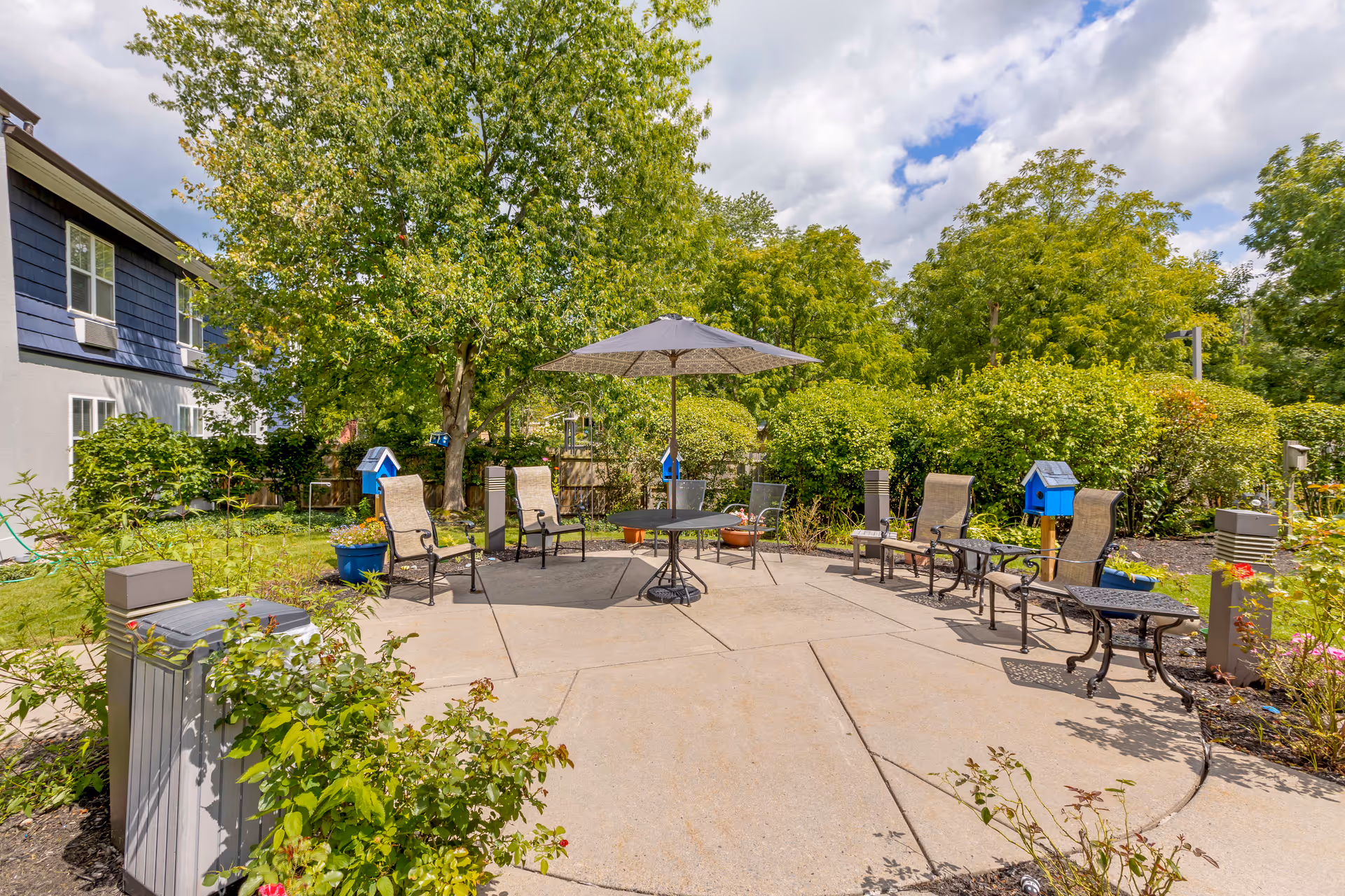 Outdoor patio area with a round table and umbrella surrounded by several chairs. The patio is bordered by green bushes and trees, with a building visible on the left side. The sky is partly cloudy.