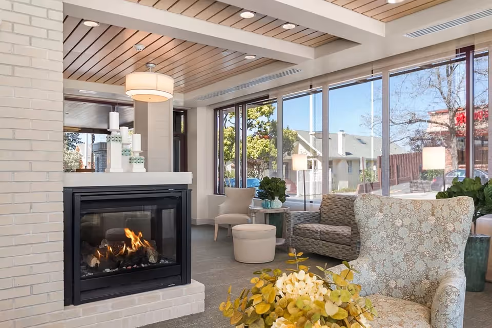 A cozy living room area with a lit fireplace surrounded by white brick. The room features large windows letting in natural light, a patterned armchair, a beige chair with an ottoman, a small round table with plants, and a sofa. The ceiling has wooden slats and recessed lighting.