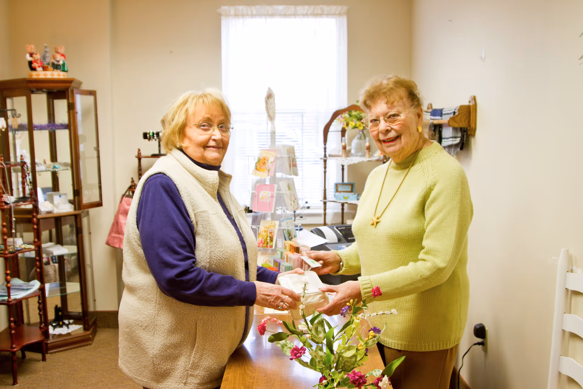 Two elderly women smiling and exchanging a small white box in a cozy room with shelves displaying various items and a window with sheer curtains in the background.
