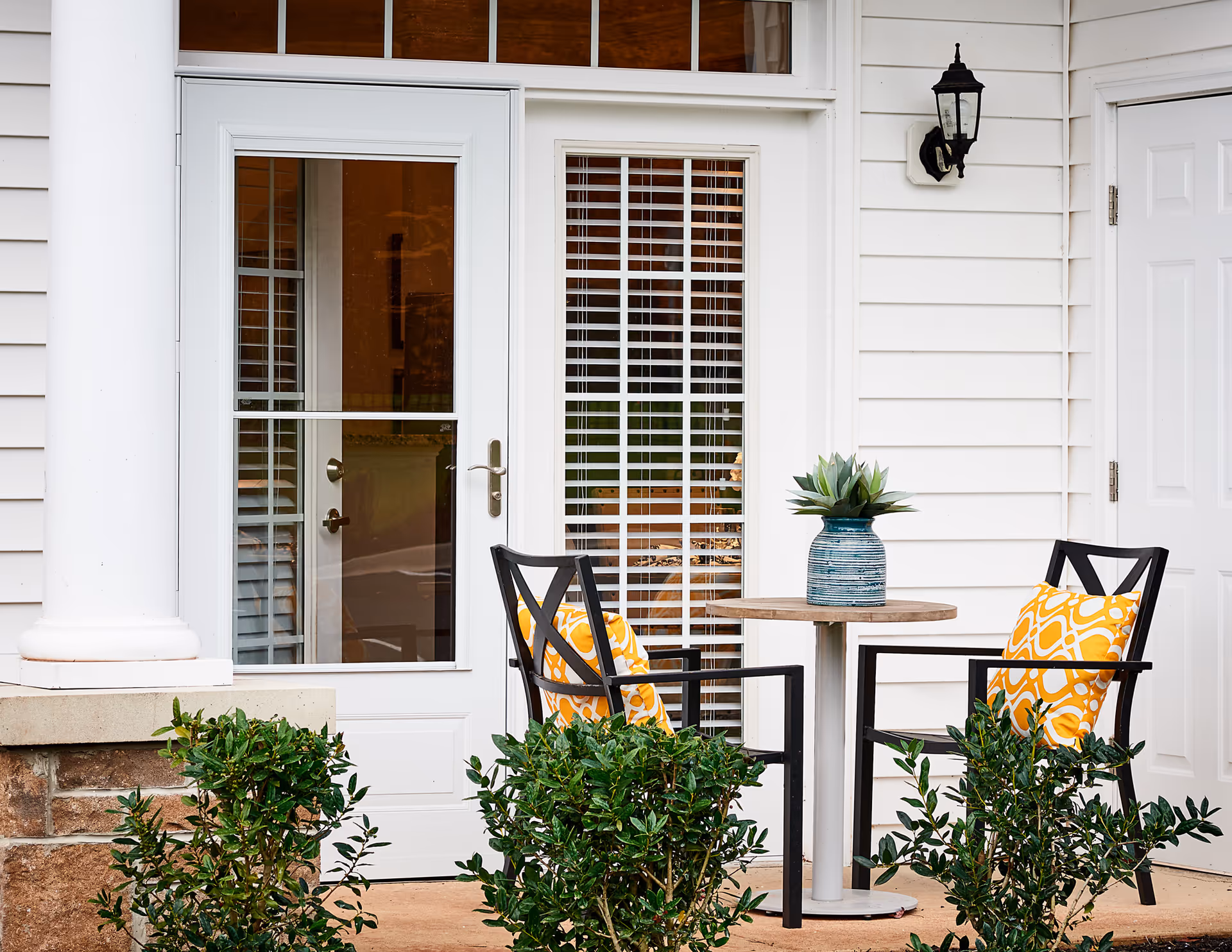 Outdoor patio area with two black chairs featuring yellow patterned cushions, a small round wooden table with a blue vase and green plant, white siding wall, white door with glass panels and blinds, and a black outdoor wall lantern.