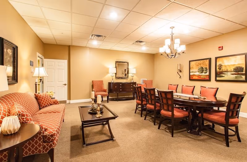 A well-lit room with beige walls and carpeted floor featuring a long dining table with eight red cushioned chairs, a chandelier above, a patterned red sofa with matching pillows, a coffee table with decorative items, two armchairs, a sideboard with lamps, and two landscape paintings on the wall.