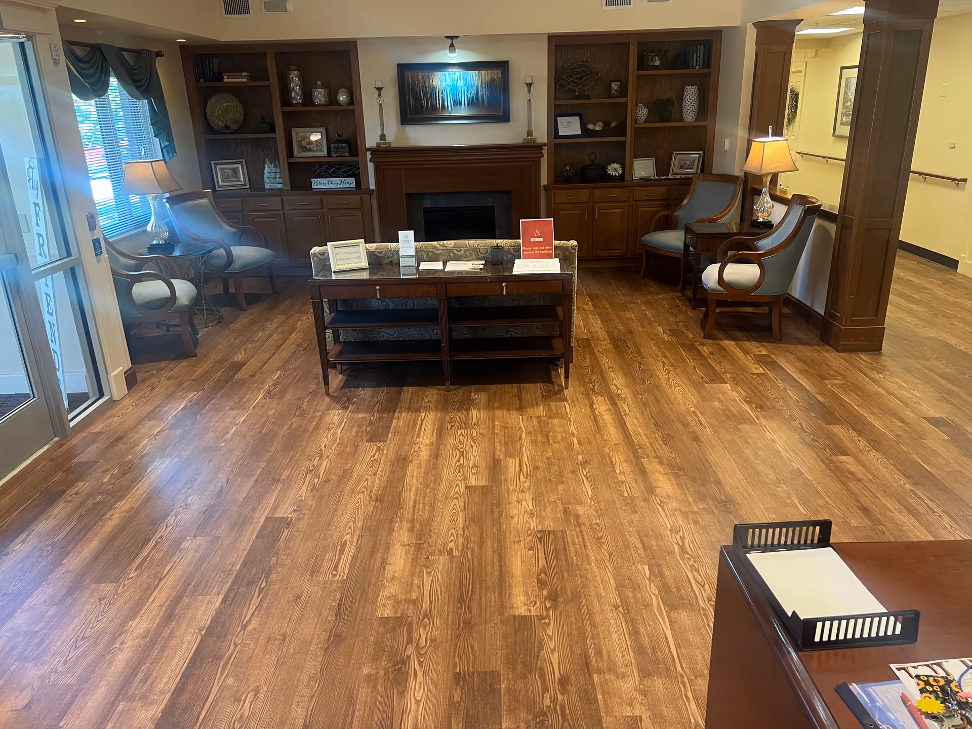A cozy senior living facility common area with wooden flooring, a fireplace centered between two built-in wooden bookshelves, and several cushioned armchairs arranged around a small table. There are lamps on side tables and a large window with curtains letting in natural light.