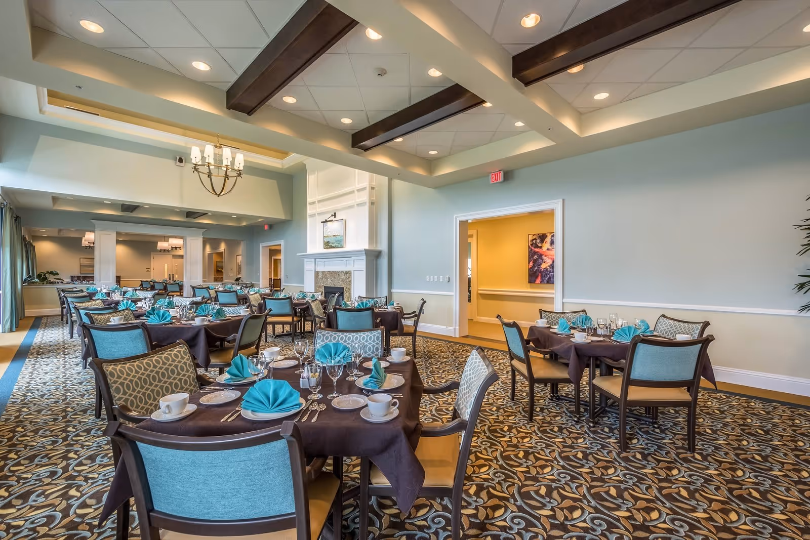 A spacious dining room with multiple round tables covered with dark tablecloths, each set with white plates, cups, silverware, wine glasses, and teal folded napkins. The room features patterned carpet, light blue walls, a white fireplace, and a ceiling with recessed lighting and wooden beams. There is a chandelier hanging from the ceiling and an exit sign above a door leading to another room.