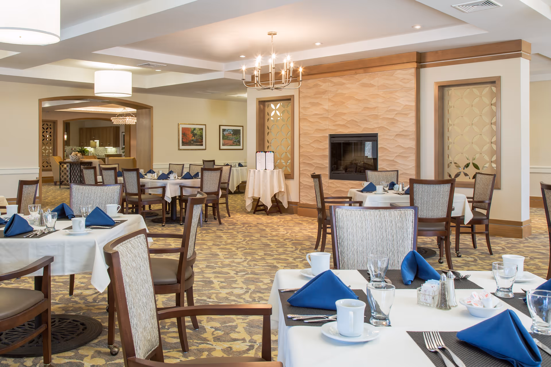 A well-lit dining room in a senior living facility with tables covered in white tablecloths, set with blue folded napkins, cups, glasses, and silverware. The room features patterned carpet, wooden chairs with cushioned seats, a decorative fireplace with a textured wall, and framed artwork on the walls. There is a chandelier and ceiling lights providing illumination.