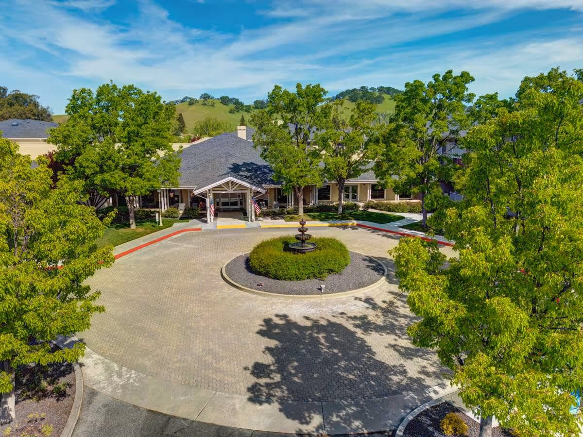 Front entrance of a senior living facility with a circular driveway, central fountain, and trees.