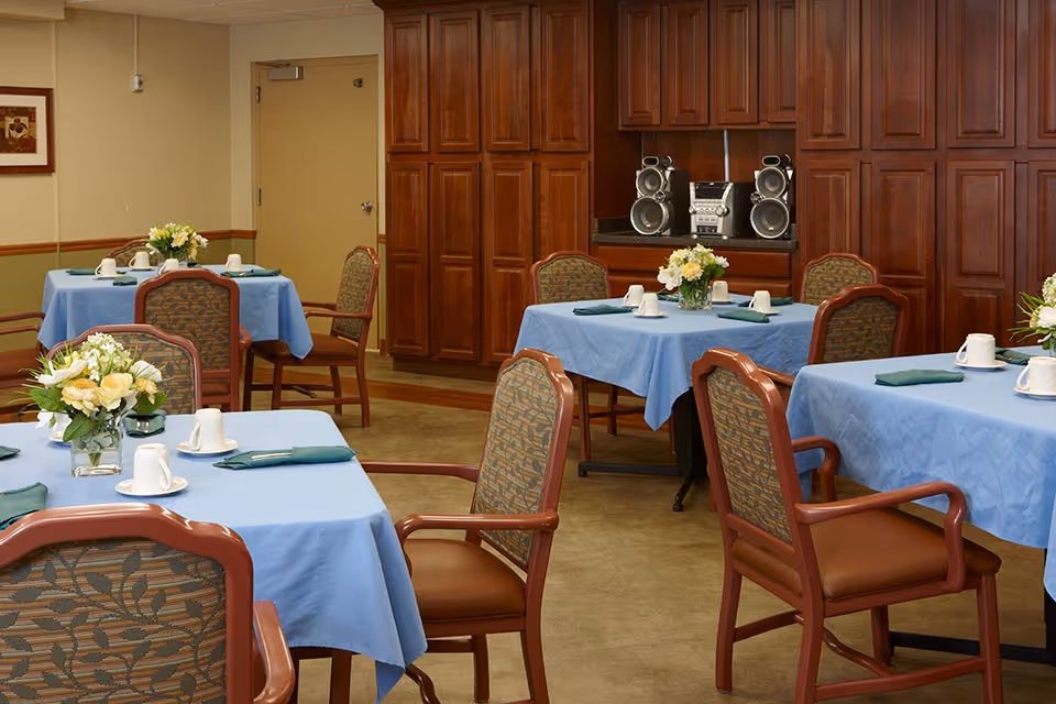 Dining room with several tables covered in blue tablecloths, each set with white cups, green napkins, and floral centerpieces. Wooden chairs with patterned upholstery surround the tables. In the background, there are wooden cabinets and a stereo system.