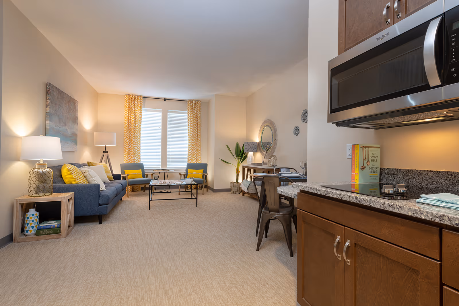 A bright and cozy living area in a senior living facility with a blue sofa, two blue chairs with yellow cushions, a coffee table, and two floor lamps. The room has large windows with yellow patterned curtains. To the right, there is a small dining area with a table and chairs, and a kitchen counter with a microwave and cookbooks on it.