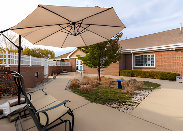 Outdoor courtyard with a large patio umbrella, lounge chairs, walkways, and landscaped beds in front of a brick building.