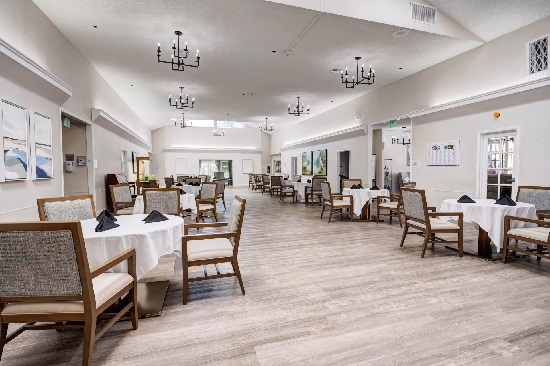 A spacious dining room with multiple round tables covered in white tablecloths and set with black folded napkins. Each table is surrounded by wooden chairs with cushioned seats and backs. The room has light-colored wooden flooring, white walls, and several black chandeliers hanging from the ceiling. There are paintings on the walls and large windows letting in natural light.