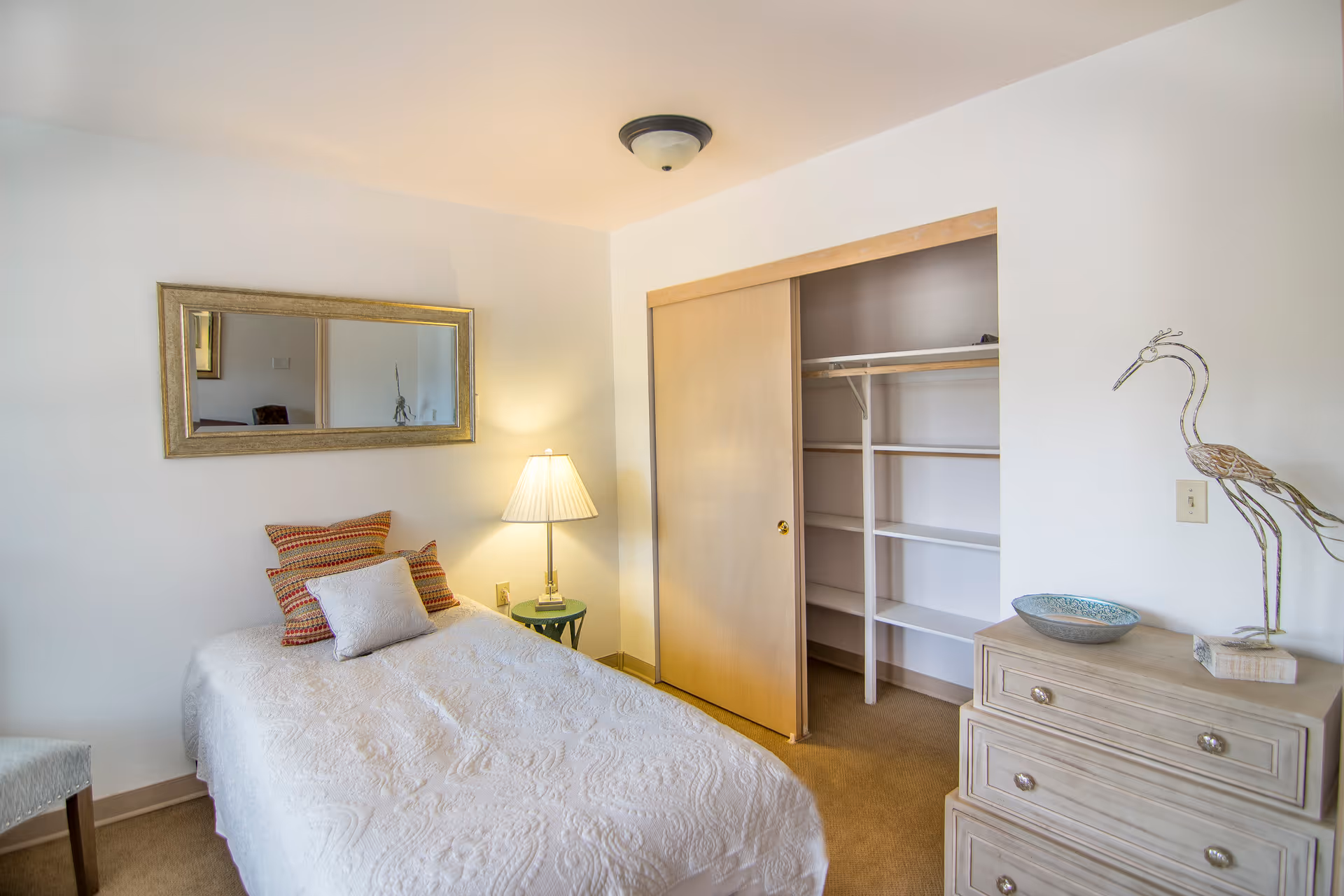 A cozy bedroom with a single bed covered in a white quilt and three decorative pillows. A small green side table with a lamp is next to the bed. There is a large rectangular mirror on the wall above the bed. To the right, there is an open closet with sliding wooden doors and built-in shelves. A light-colored dresser with a decorative bird sculpture and a bowl is also visible. The walls are white and the carpet is beige.