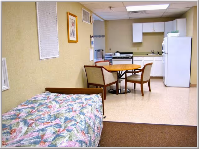 Studio-style interior with a single bed in the foreground, a round table with chairs, and a kitchenette with stove and refrigerator in the background.