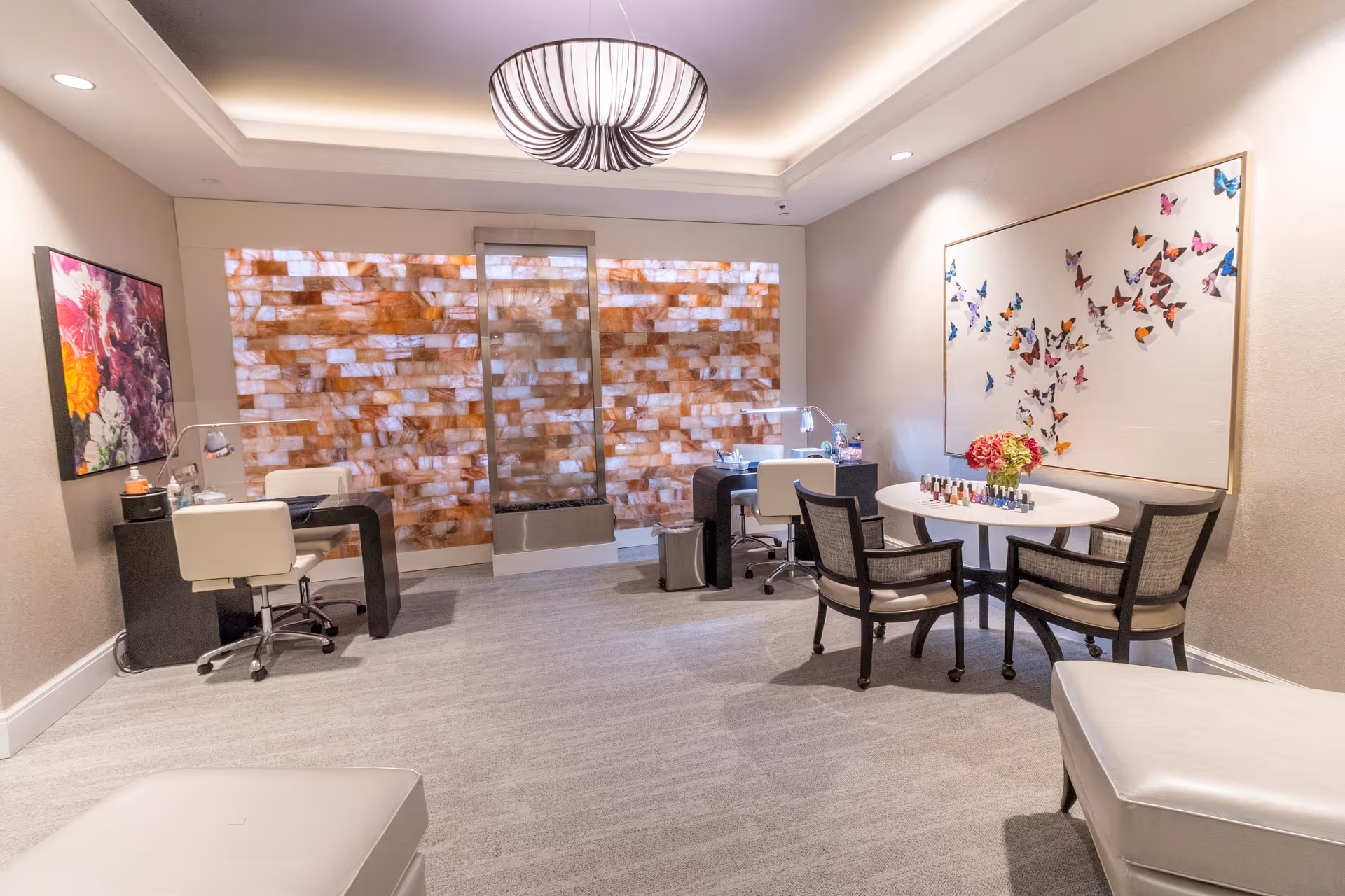 A well-lit interior room with a modern design featuring two manicure stations with white chairs and black desks, a round table with four chairs, and colorful butterfly artwork on the wall. The back wall has a decorative panel with a warm, amber-colored stone pattern, and a large, circular ceiling light fixture hangs above.