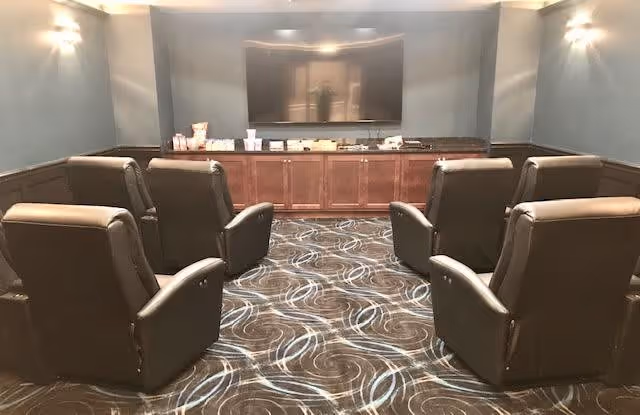 A small theater or media room with six brown leather recliner chairs arranged in two rows facing a large flat-screen TV mounted on a gray wall. Below the TV is a wooden cabinet with snacks and drinks placed on top. The room has patterned carpet and wall sconces providing soft lighting.