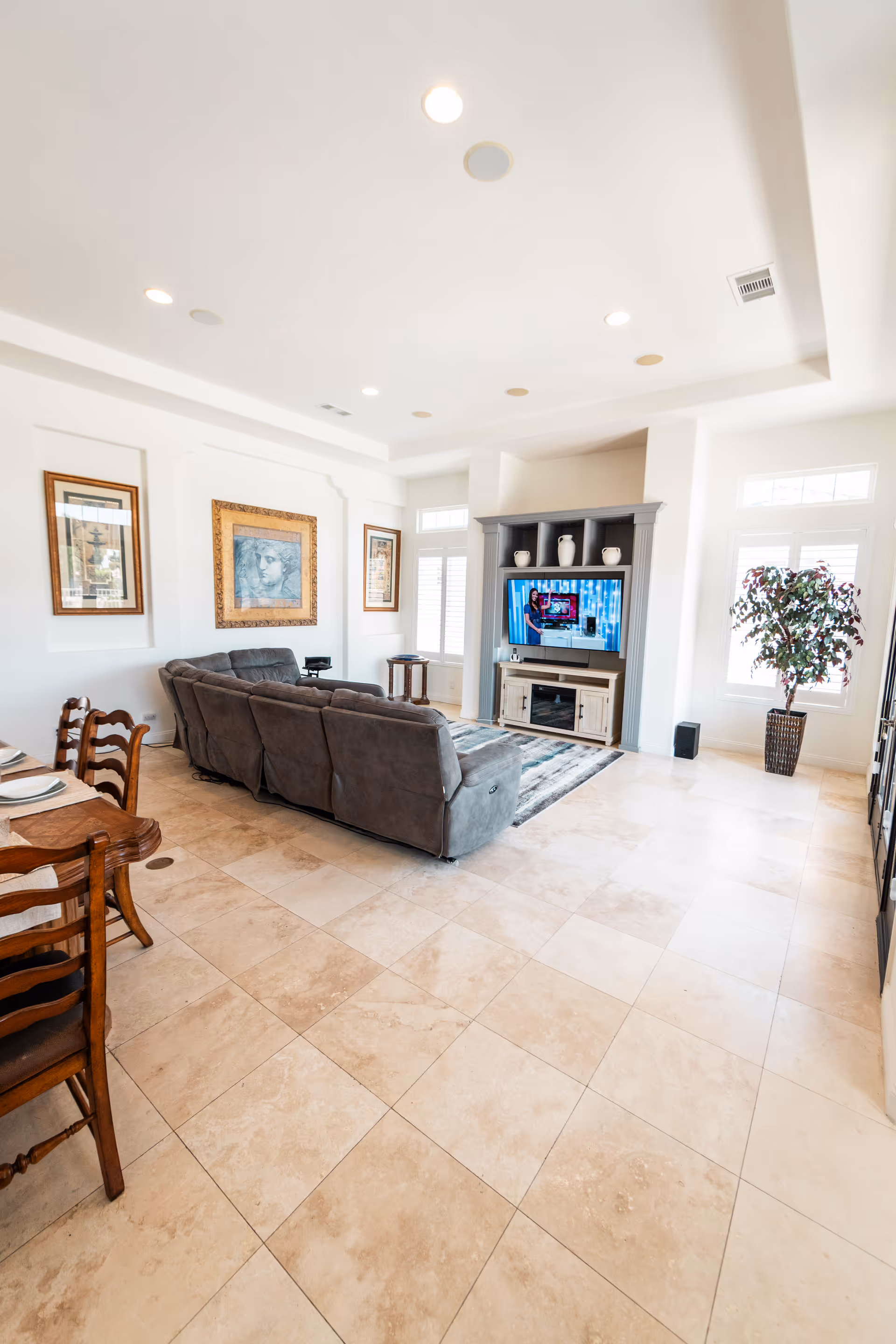 A bright living room with beige tiled flooring, a gray sectional sofa facing a TV mounted on a gray entertainment center. The room has white walls with three framed artworks, large windows with white shutters, a potted plant near the window, and a wooden dining table with chairs on the left side.