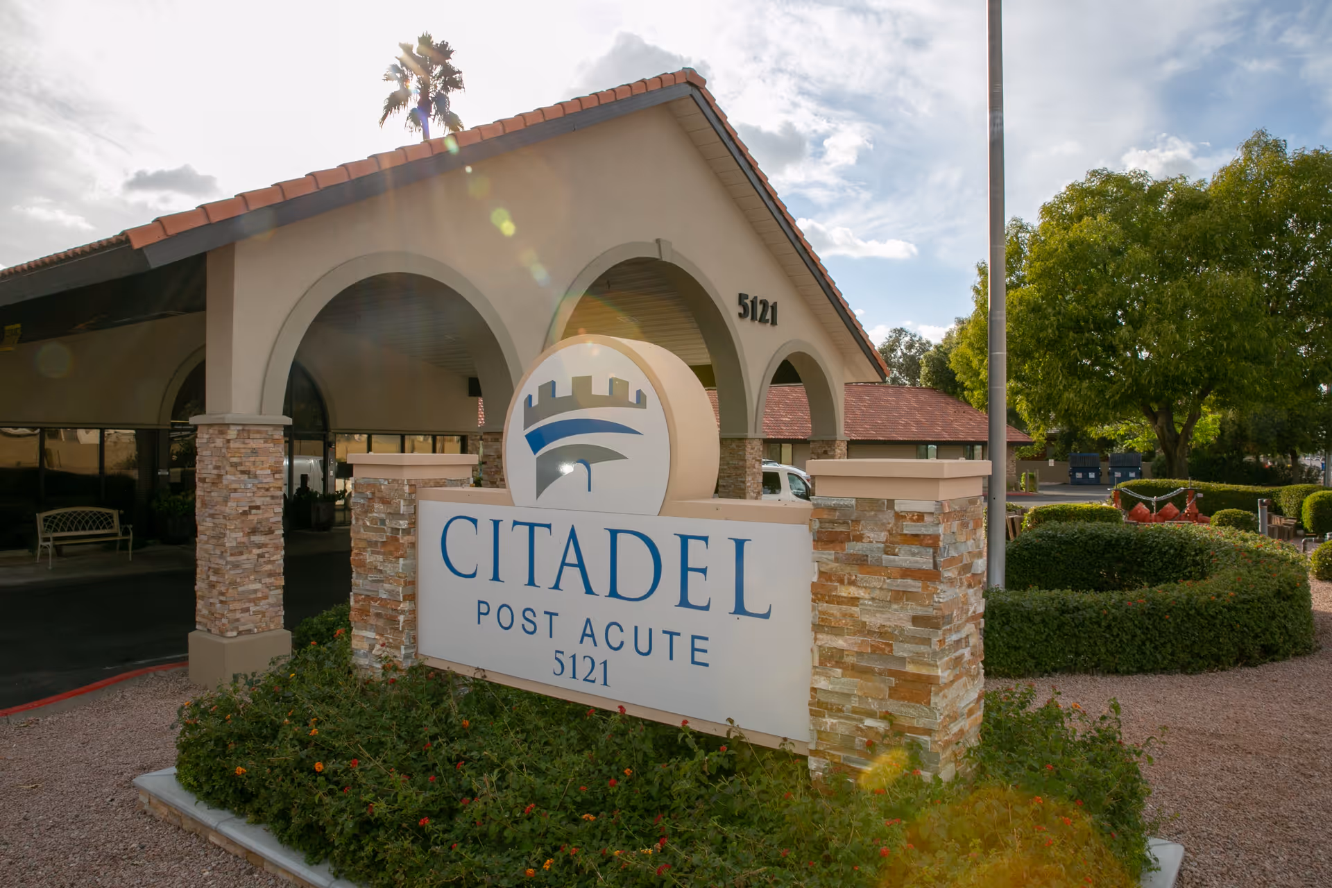Exterior view of Citadel Post Acute facility with a stone and stucco sign displaying the name and address 5121, surrounded by greenery and landscaping. The building has a tiled roof and arched entrance.