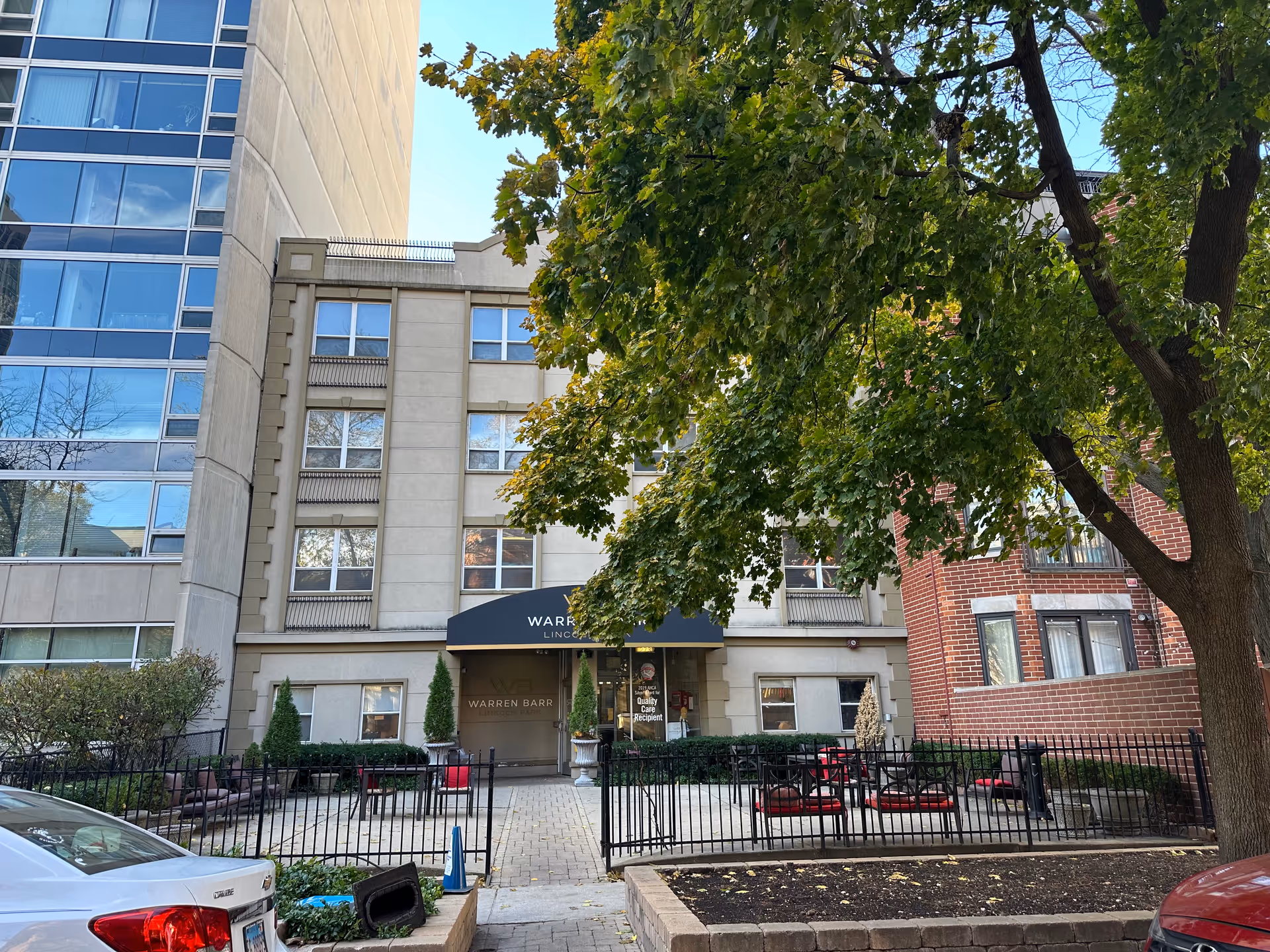 Exterior view of the Warren Barr Lincoln Park facility entrance with a black awning displaying the facility name. The building is surrounded by trees, outdoor seating with red cushions, and a black metal fence. Cars are parked in front of the building.