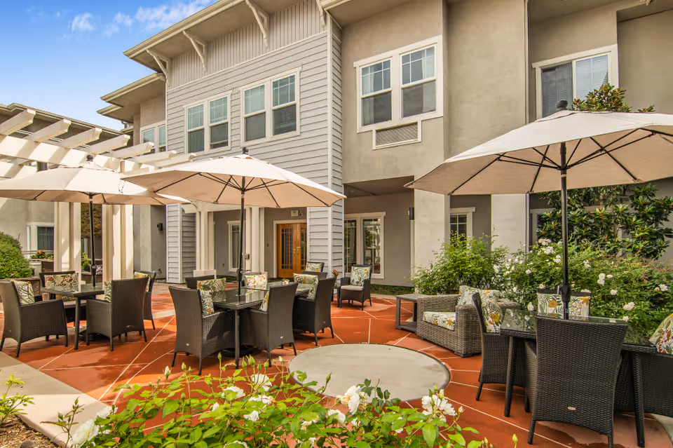 Outdoor courtyard patio with umbrella-shaded tables, wicker chairs, and seating in front of a multi-story residence.