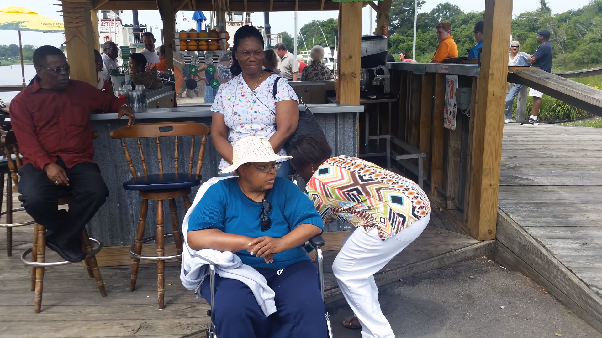 A group of people gathered outdoors near a wooden structure with a counter. One woman in a wheelchair wearing a white hat and blue shirt is in the foreground, while another woman in a colorful patterned shirt is bending down to speak to her. Other people are standing or sitting around the counter, with trees and water visible in the background.