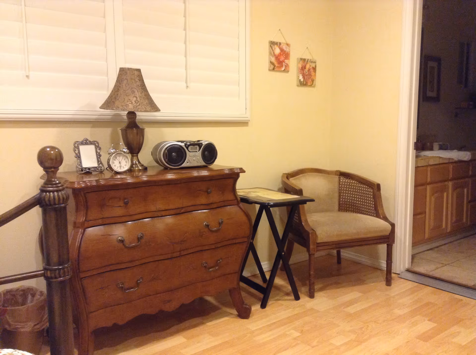 A cozy interior corner featuring a wooden dresser with a decorative lamp, an alarm clock, a small framed picture, and a radio on top. Next to the dresser is a small folding table and a cushioned wooden chair. The room has light-colored walls with two floral wall hangings and light wood flooring. A doorway leads to a kitchen area with wooden cabinets and a tiled floor.