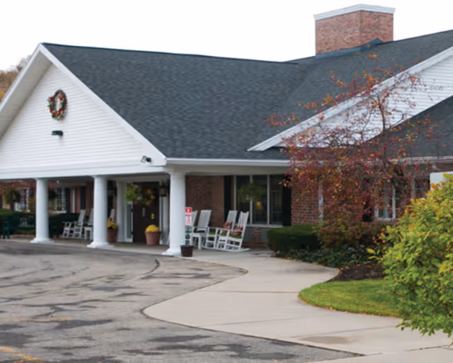 Front entrance of a brick senior living building with a covered portico supported by white columns, rocking chairs on the porch, and a curved driveway and walkway.
