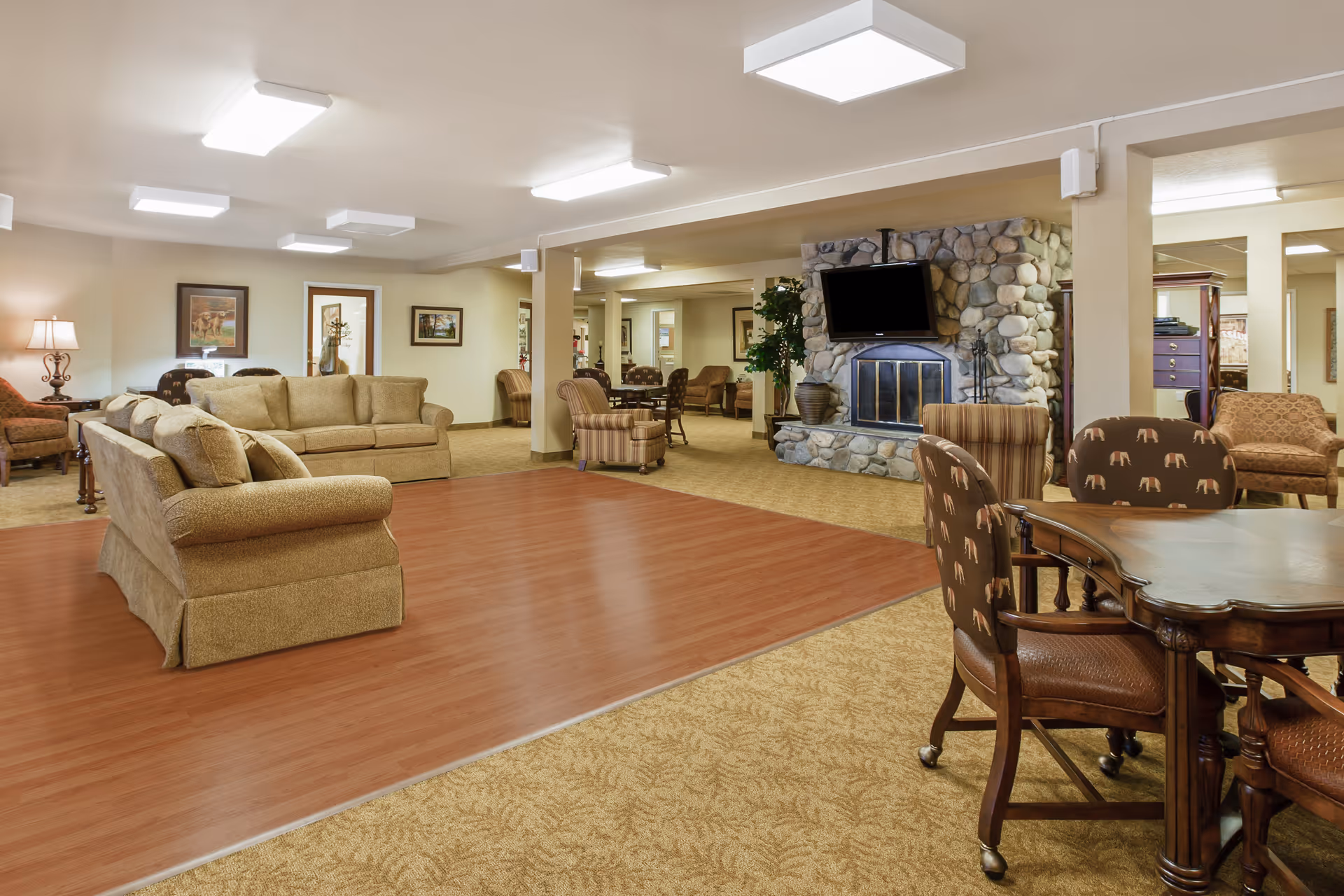 A spacious living room area in a senior living facility featuring beige sofas, patterned armchairs, a stone fireplace with a mounted flat-screen TV, wooden tables with chairs, framed artwork on the walls, and bright ceiling lights.
