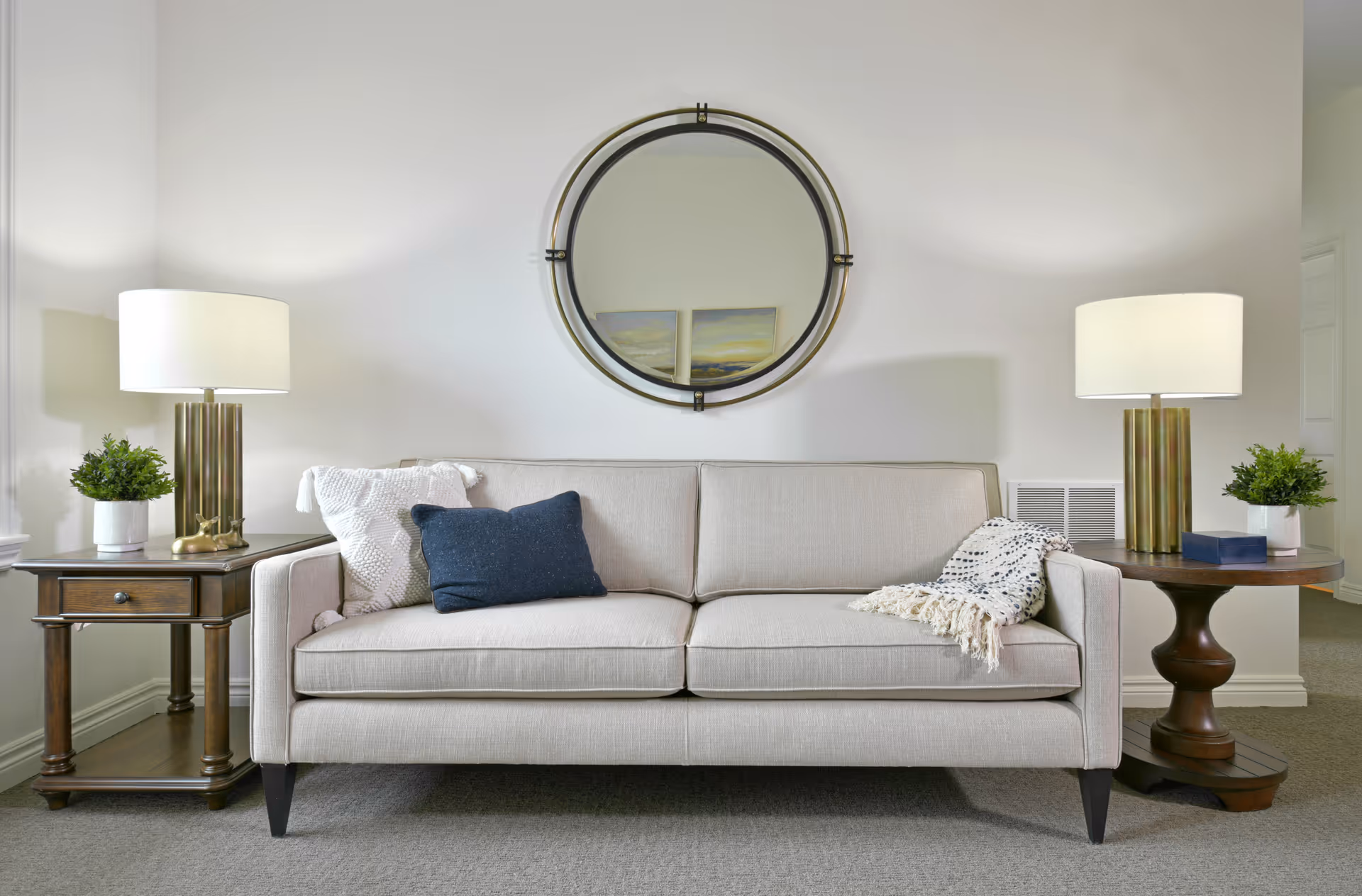 A beige sofa with two cushions and a throw blanket is centered between two wooden side tables, each with a lamp and a small potted plant. A round mirror hangs on the wall above the sofa, reflecting part of a painting.