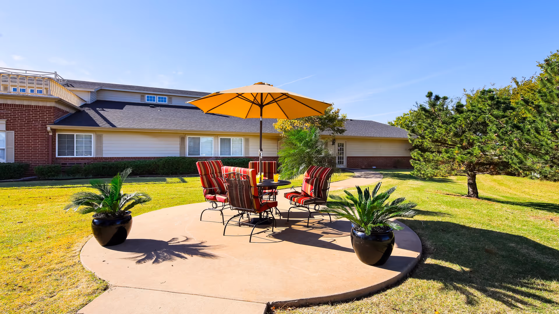 Outdoor patio area with a round concrete platform featuring a table with a yellow umbrella and four red cushioned chairs. Two large potted plants flank the seating area. The background shows a single-story building with brick and siding exterior, windows, and a door, under a clear blue sky.