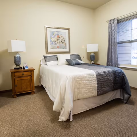 Furnished bedroom with a double bed, two bedside tables and lamps, framed artwork above the bed, and a window with curtains.