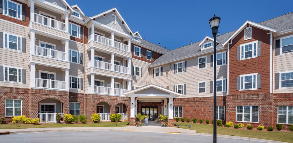 Exterior view of a multi-story senior living facility building with a combination of brick and beige siding, white balconies, and a covered entrance. The building is surrounded by neatly trimmed bushes and a clear blue sky.