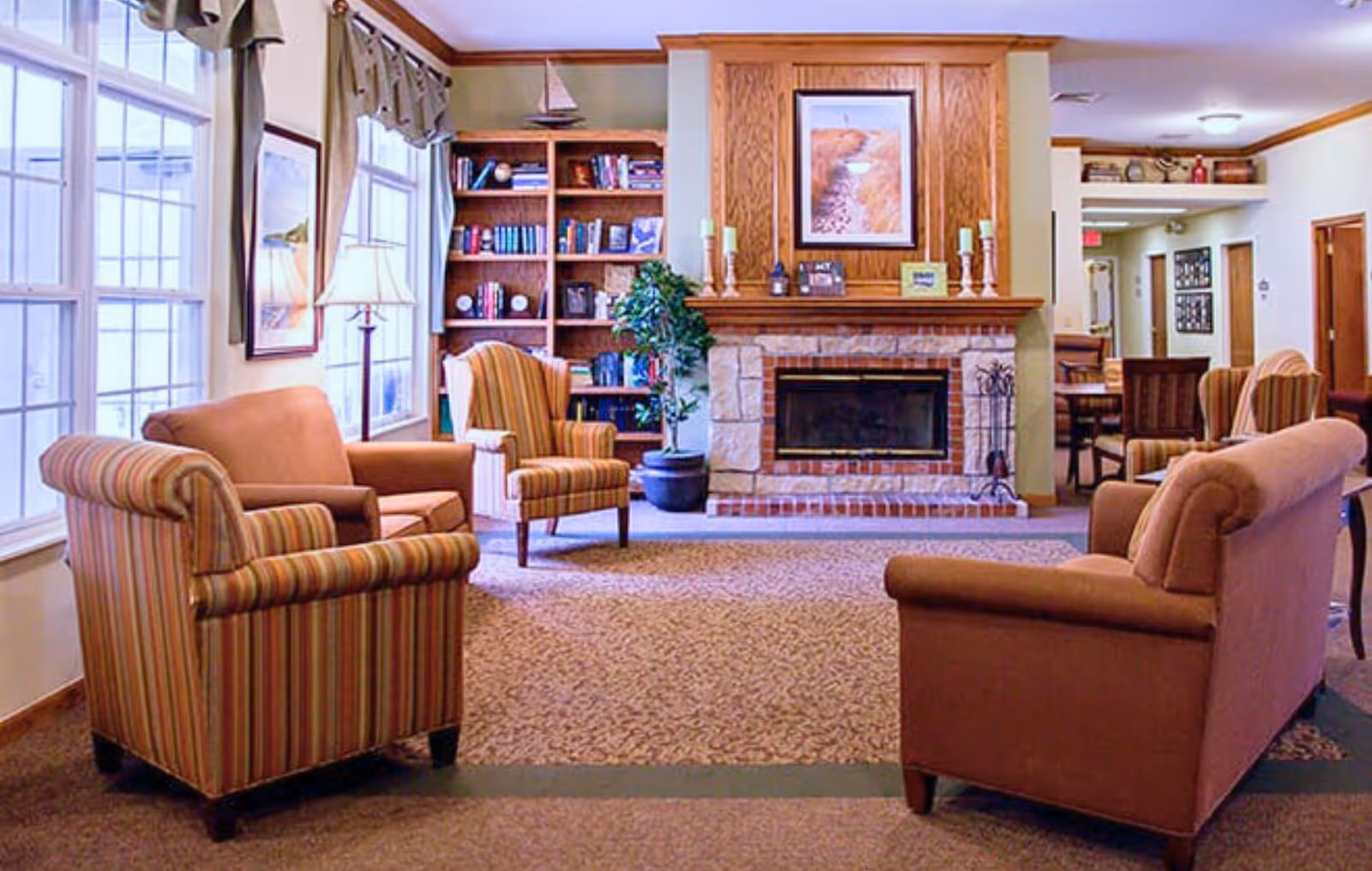 A cozy living room area with a brick fireplace centered on the back wall, flanked by wooden bookshelves filled with books. The room has large windows on the left side with curtains, and is furnished with striped and solid upholstered armchairs and sofas arranged around the fireplace. There is a framed picture above the fireplace and decorative candles on the mantel. The carpeted floor and warm lighting create a welcoming atmosphere.