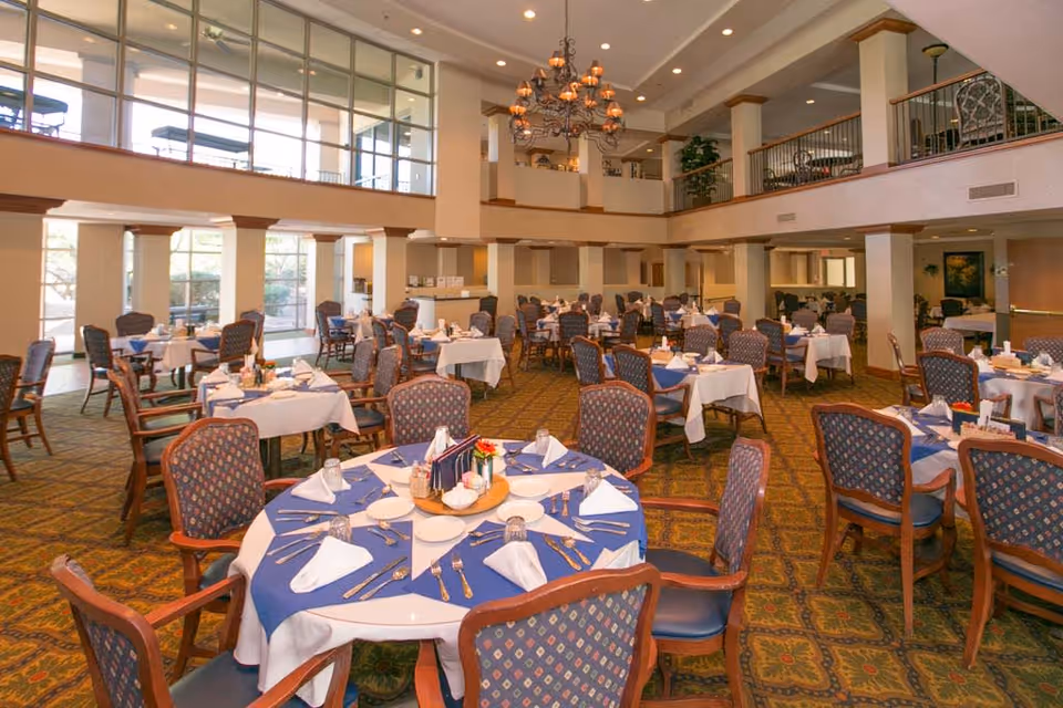 Spacious senior dining room with round tables set with blue tablecloths and chairs beneath a mezzanine and chandelier.