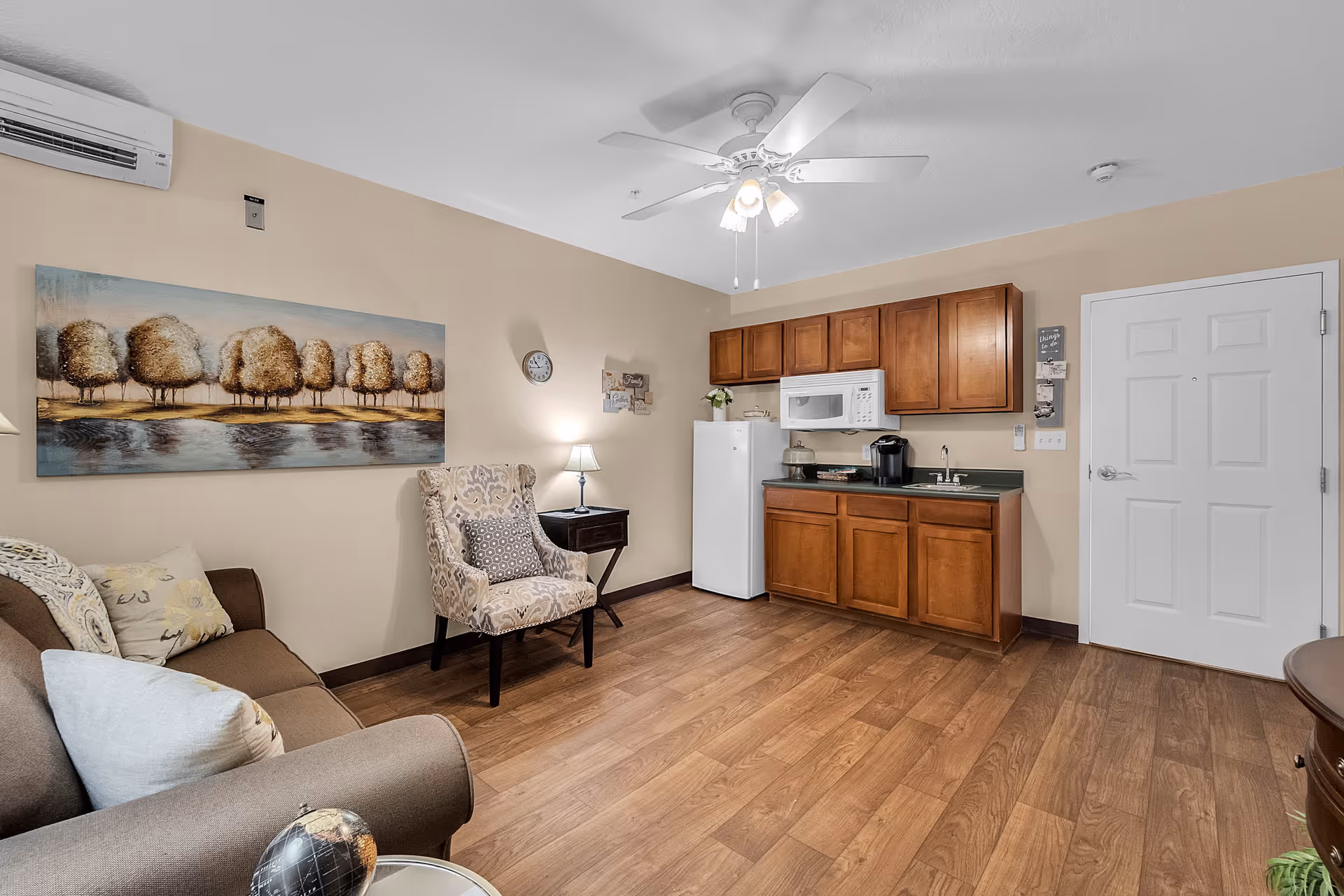 A cozy living area in The Grande featuring a brown sofa with pillows, a patterned armchair, a small side table with a lamp, and a kitchenette with wooden cabinets, a white refrigerator, microwave, and coffee maker. The room has wood flooring, beige walls, a ceiling fan with lights, and a large painting of trees over a body of water on the wall.
