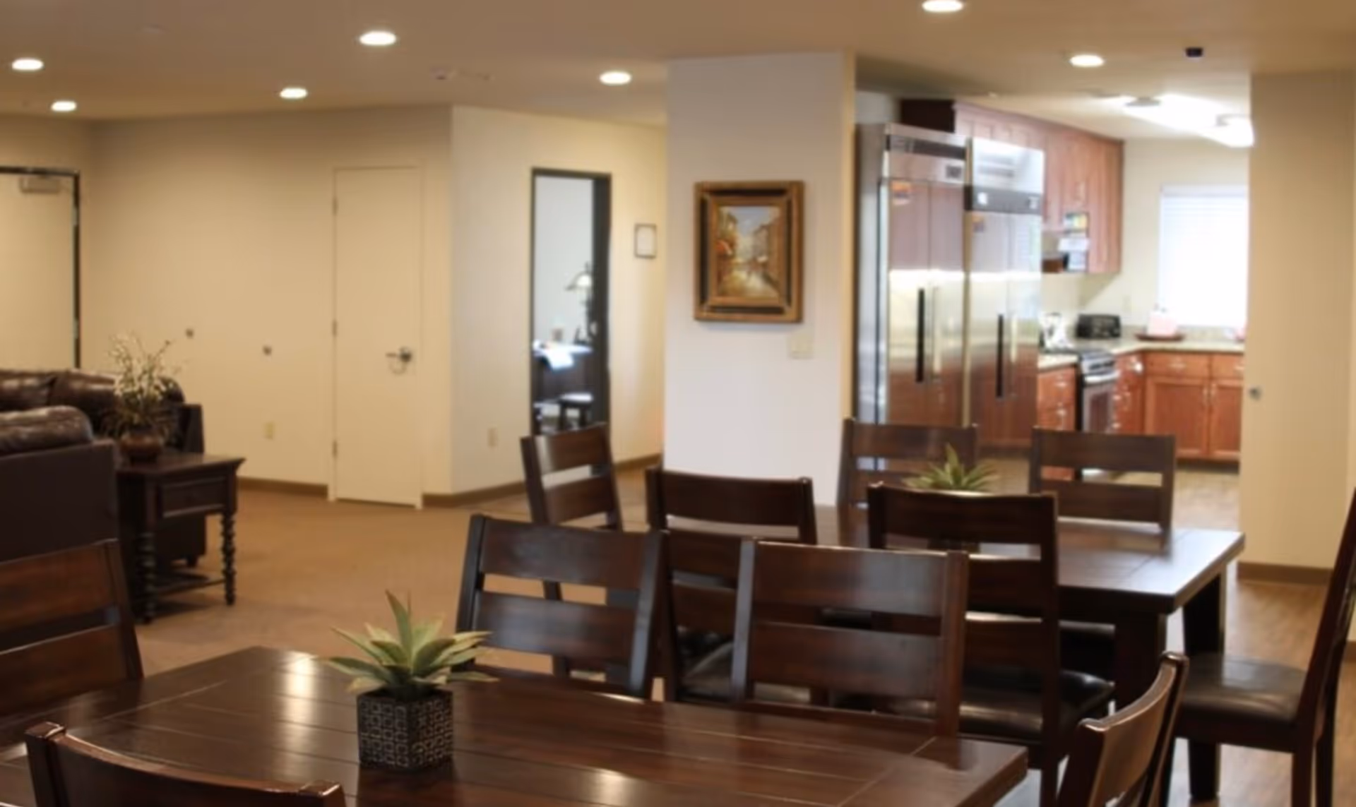 Interior view of a senior living facility showing a dining area with dark wooden tables and chairs, a small potted plant on the table, a kitchen with stainless steel appliances and wooden cabinets in the background, and a living area with leather sofas and a side table with a plant.