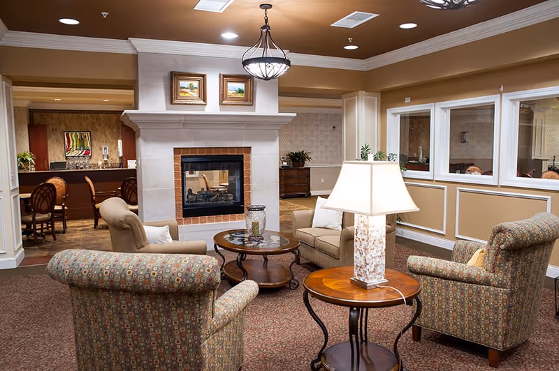 Cozy lounge with patterned armchairs and side tables arranged around a central fireplace in a senior living facility.
