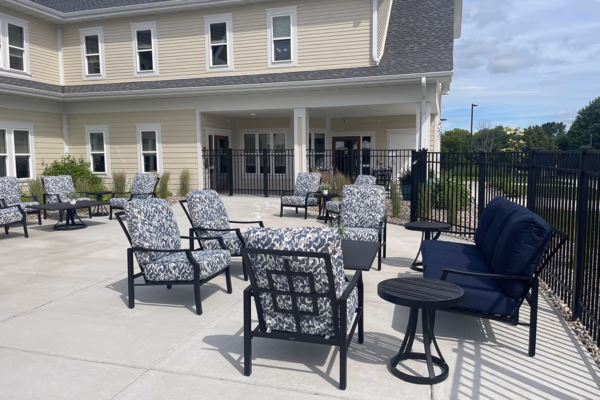 Outdoor patio area at Volante Senior Living of Hanover with multiple cushioned chairs and small tables arranged on a concrete surface next to a beige building with white trim and several windows. The area is enclosed by a black metal fence and there is greenery and a clear sky in the background.