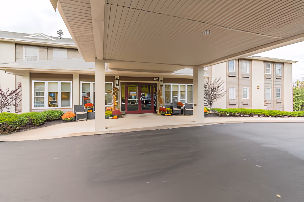 Covered entrance area of a senior living facility with seating arrangements and potted plants near the glass double doors. The building exterior is beige with multiple windows and some greenery around the entrance.