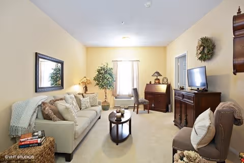 A cozy living room with a beige sofa adorned with pillows and a throw blanket, a wooden coffee table with decorative items, a large mirror on the wall, a potted plant near a window with curtains, a wooden desk with a chair, a TV on a wooden cabinet, and an upholstered chair with a pillow. The walls are painted light yellow and the room is well-lit with natural light.