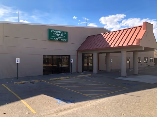 Exterior view of a single-story medical facility building with a beige facade and a red metal awning over the entrance. A green sign above the windows reads 'Community Hospital and St. Marys Clinic.' There are several handicap parking spaces in front of the building under a clear blue sky with some clouds.