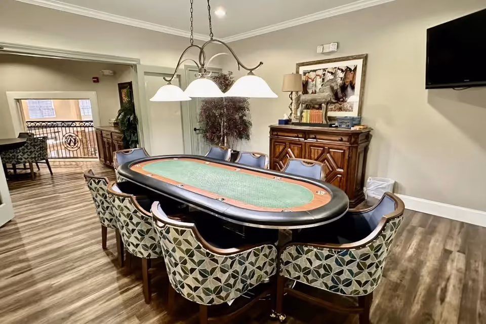 A well-lit room with a large oval poker table surrounded by eight patterned chairs. Above the table hangs a chandelier with three lights. In the background, there is a wooden cabinet with decorative items and a framed picture on the wall. A flat-screen TV is mounted on the right wall, and the floor is covered with wood-style flooring.