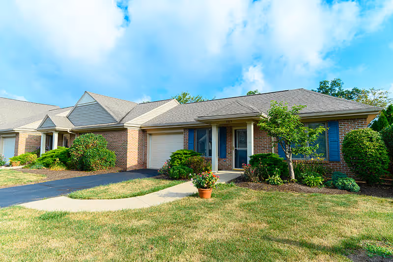 Exterior view of a single-story brick residential building with a garage, blue shutters, a small porch, and well-maintained landscaping including bushes, a small tree, and a potted plant on the lawn under a partly cloudy sky.